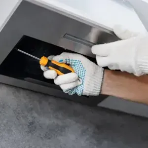 Gloved hand cleaning a vent with a brush under a white appliance.
