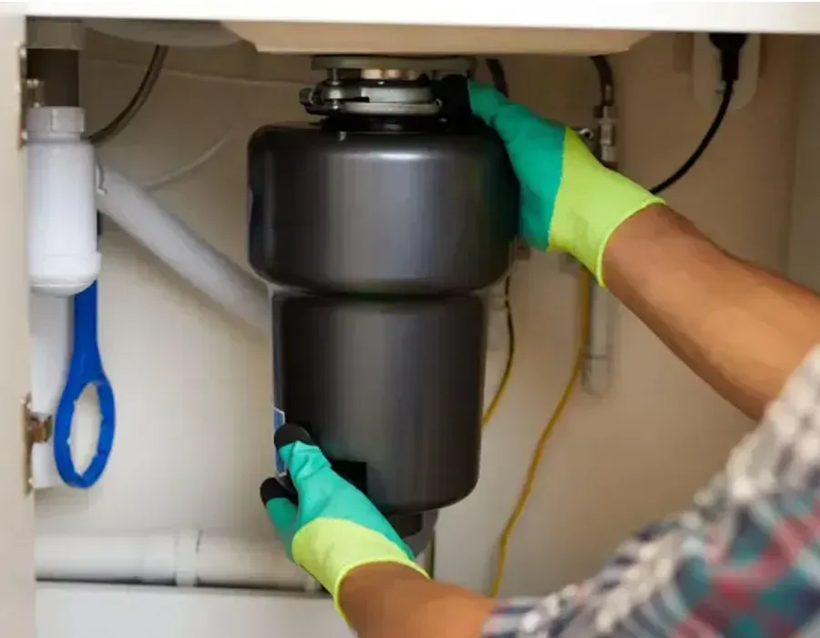 Person in green gloves installing a black garbage disposal under a kitchen sink