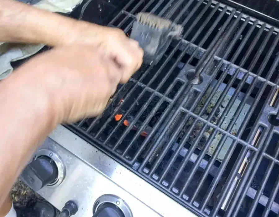 Hands cleaning a black gas grill with a brush while orange flames glow beneath the grates