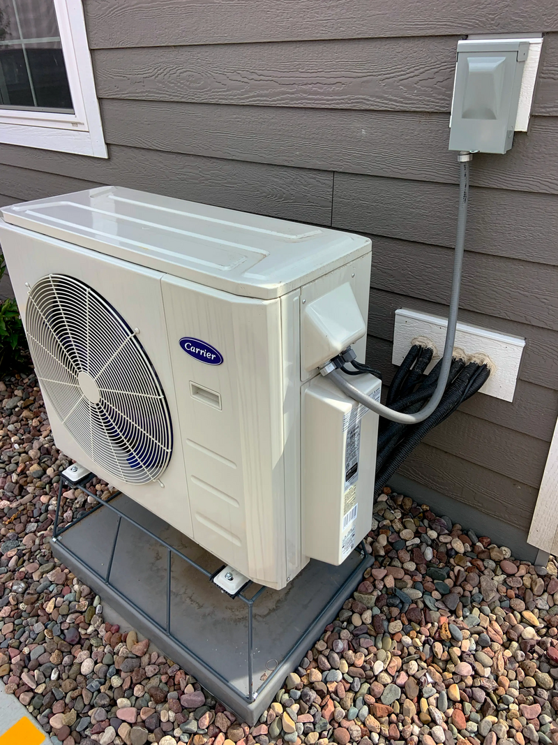 An air conditioning unit is mounted outside on a gray platform against a gray wall with electrical components attached.