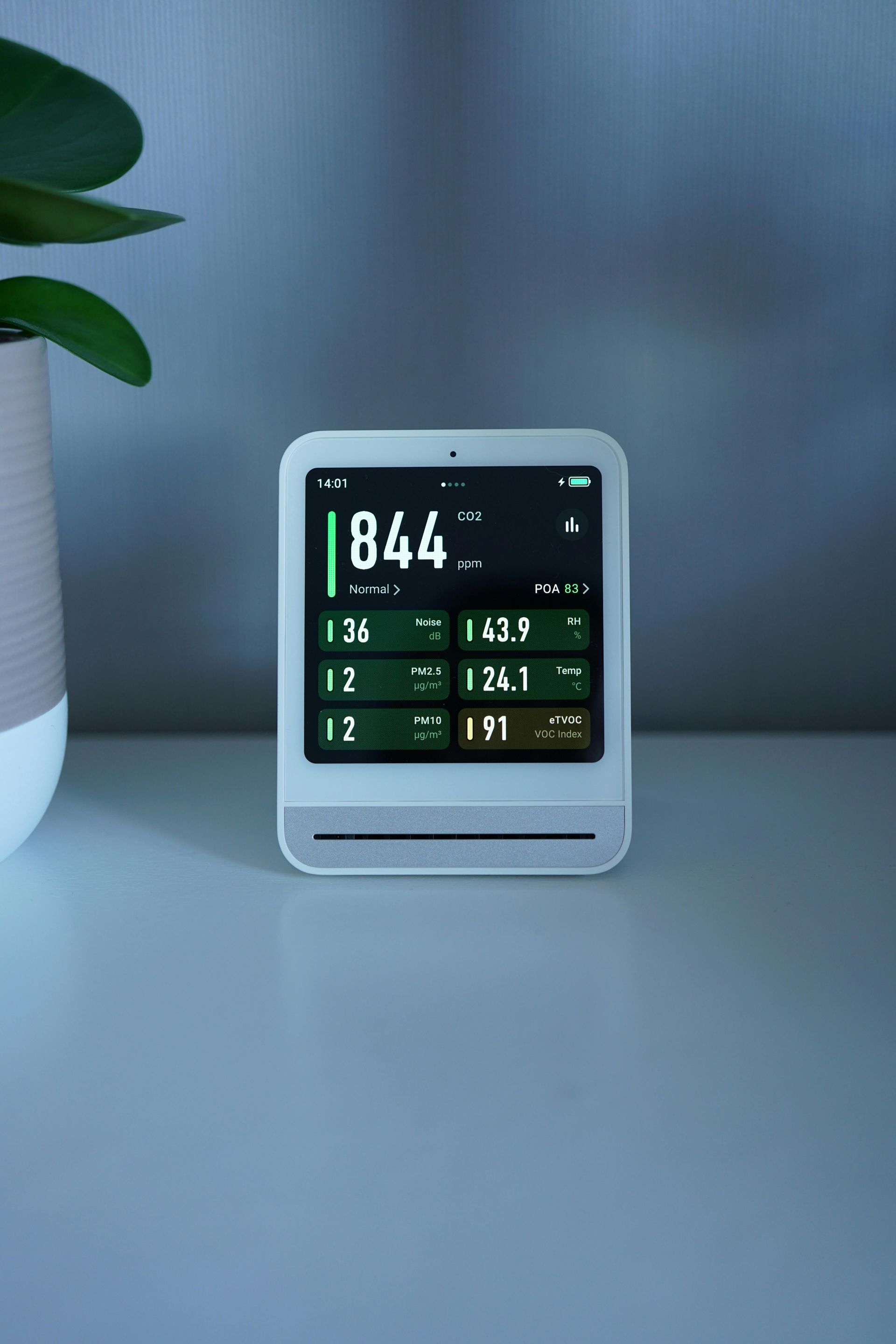 White air quality monitor displaying readings on a nightstand with a potted plant in the background.