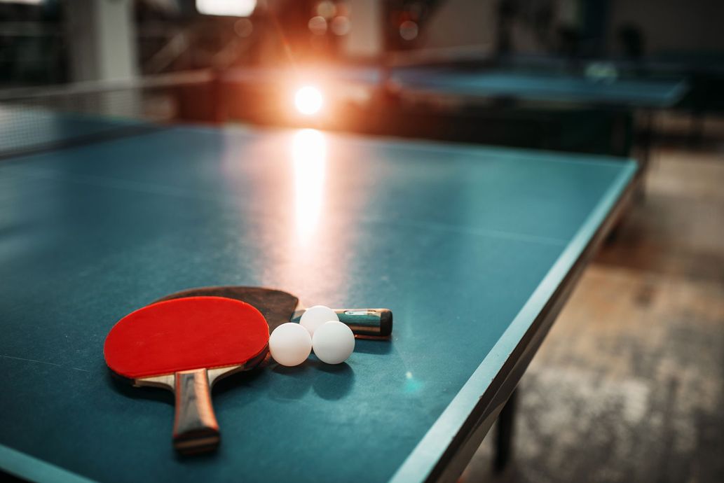 Red ping-pong paddle and white balls on a blue table tennis table, with sunlight in the background