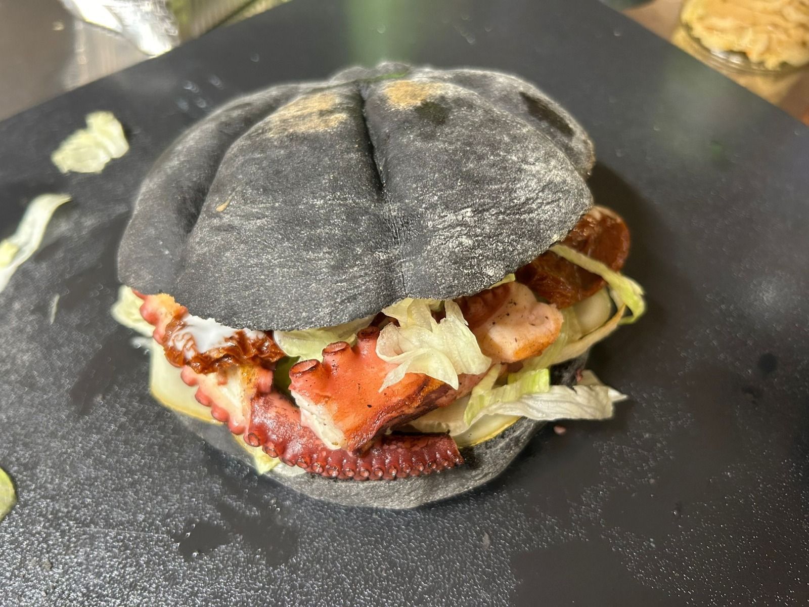 Panino nero ripieno di polpo e lattuga su una superficie nera.