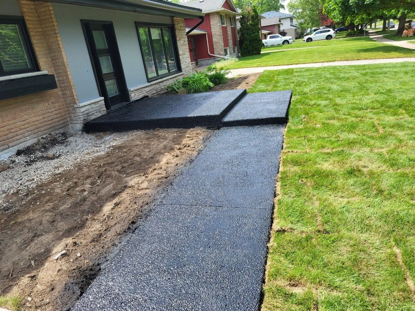 Newly installed asphalt walkway and front entry pad beside a brick home with exposed soil