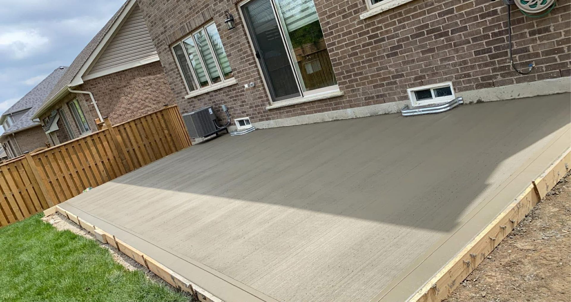 A large, freshly poured concrete shed pad sits flush against a dark brick house, featuring a smooth surface with a neatly tooled border around the edges