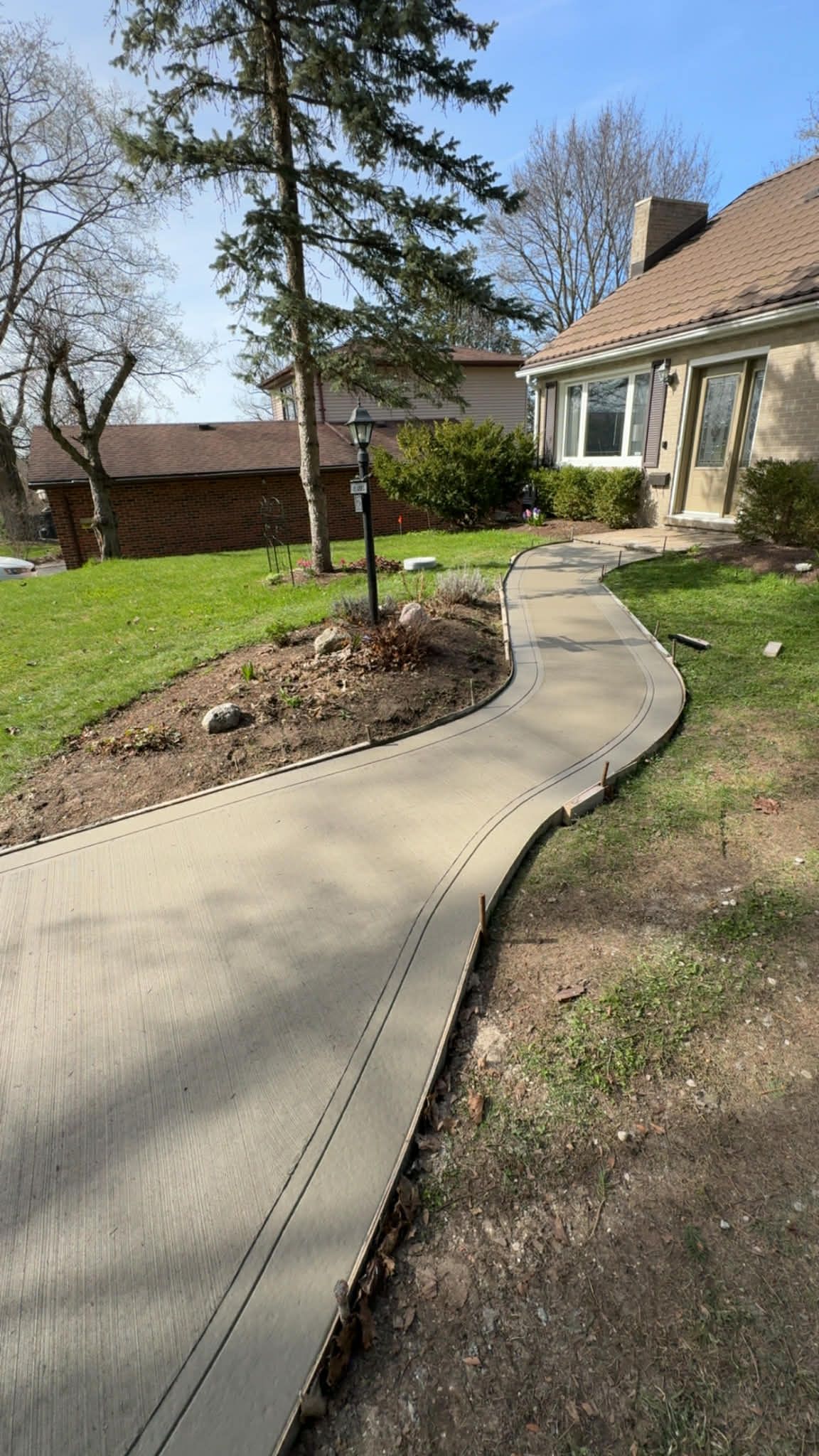 Curved concrete walkway beside a house, bordered by grass and a small landscaped tree under a blue sky.