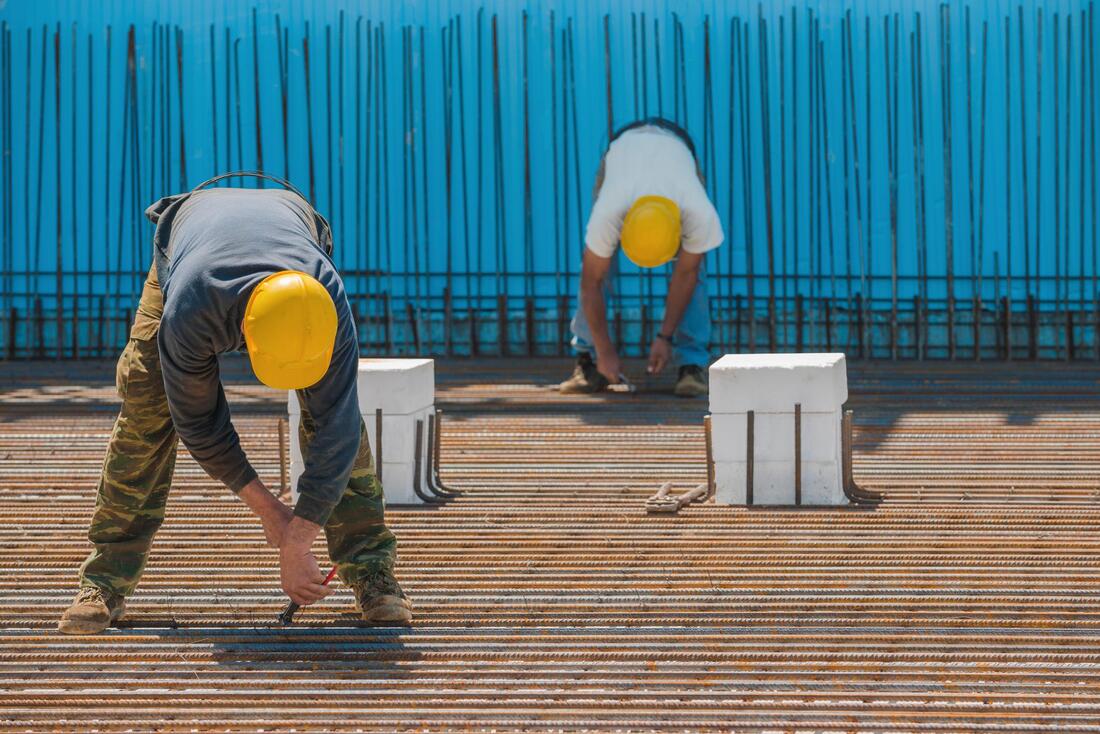 Construction workers bending over rebar on a rooftop, wearing yellow hard hats near a blue fence