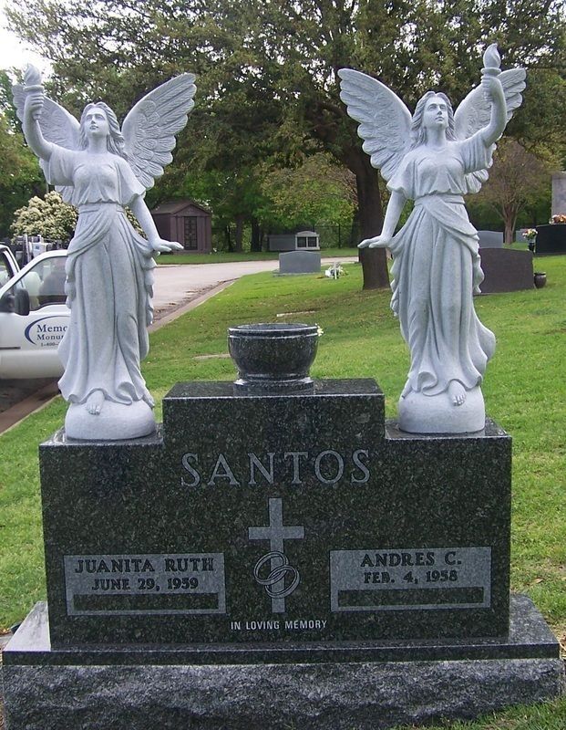 White angel statues on a dark granite grave marker in a cemetery, with the name SANTOS engraved.