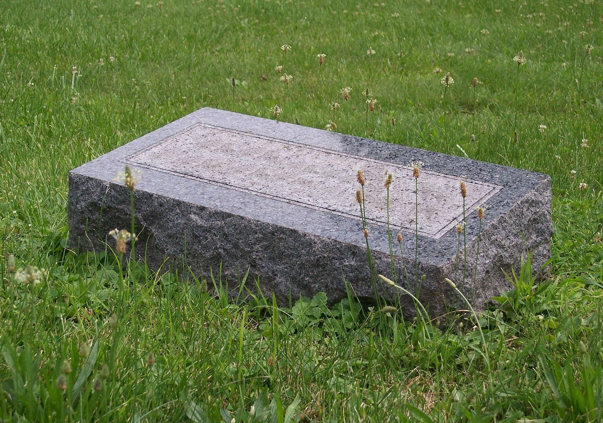 Gray stone grave marker in green grass, with a few small weeds around it