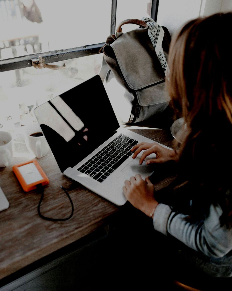 Person typing on a laptop at a desk with notebooks, cups, and an orange portable hard drive.