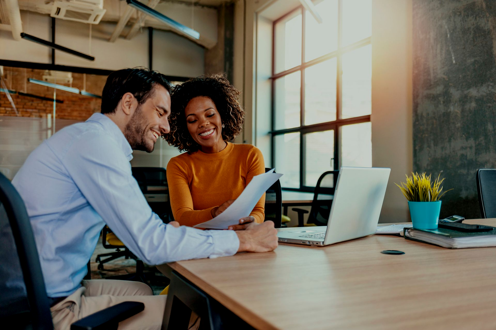 Two coworkers reviewing a document at a desk in a bright office with a laptop and plant.