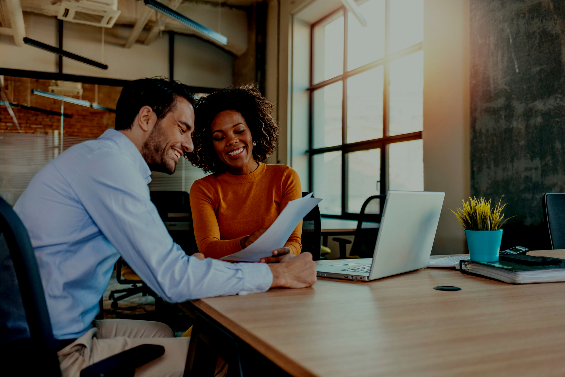 Two coworkers smiling over papers at a desk with a laptop in a bright office