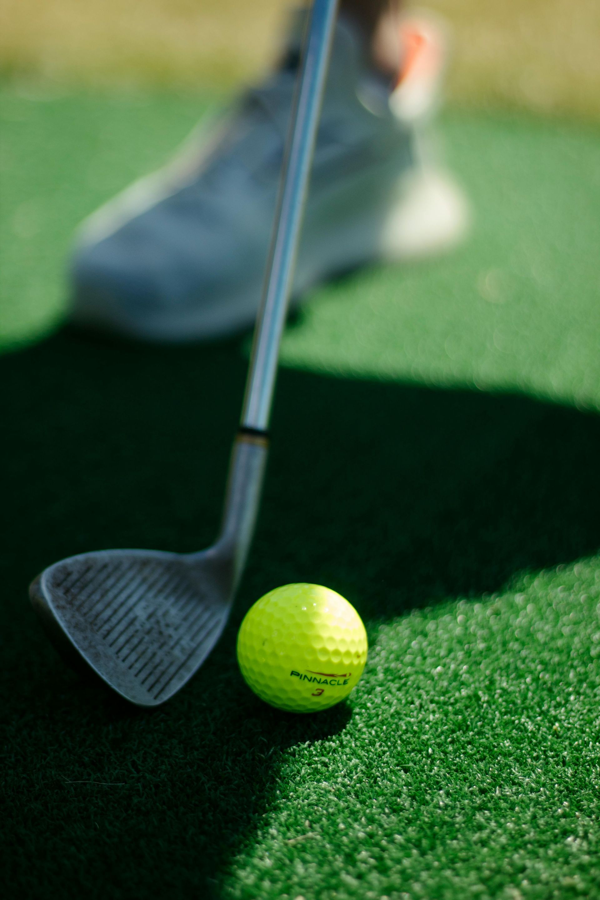 Golf club poised behind a yellow ball on green turf, with a blurred golfer in the background.