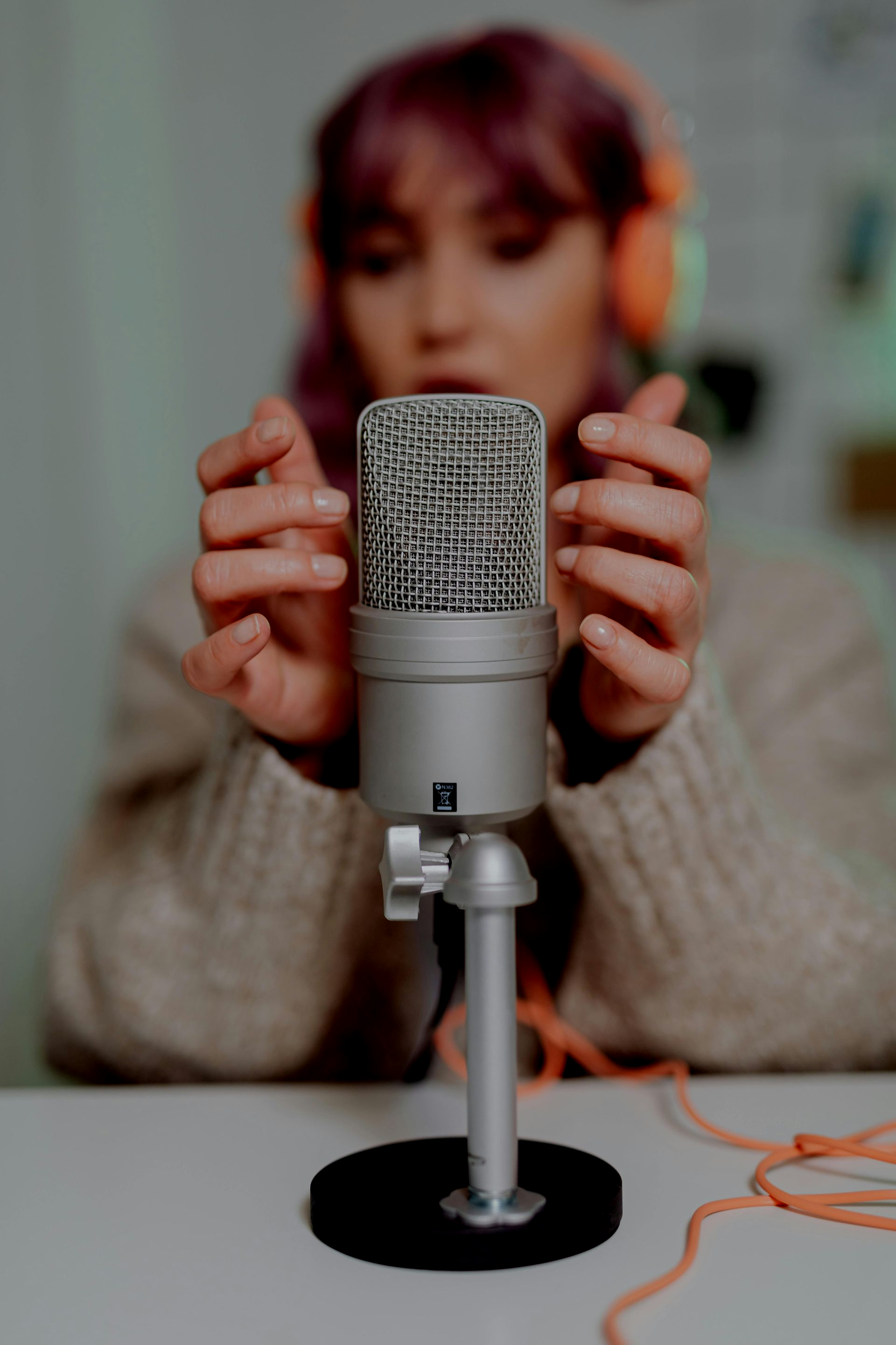 Person adjusting a silver microphone on a table, with orange headphones in the background