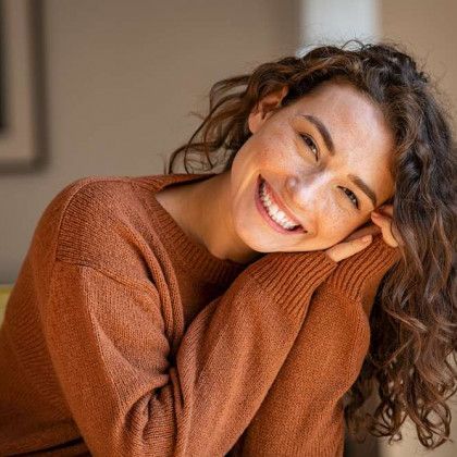 A woman in a brown sweater is smiling with her hands on her face