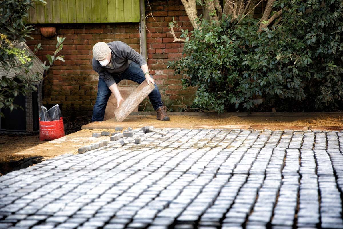 A man is laying bricks on a driveway in front of a brick wall.