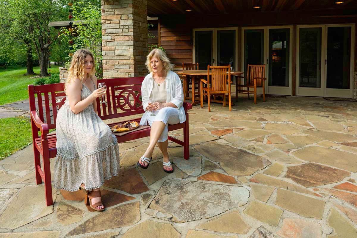 Two women sit on a bench on a hardscaped patio drinking wine
