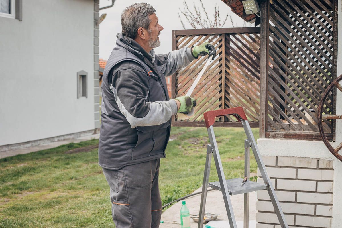 A man is standing on a ladder painting a wooden fence.