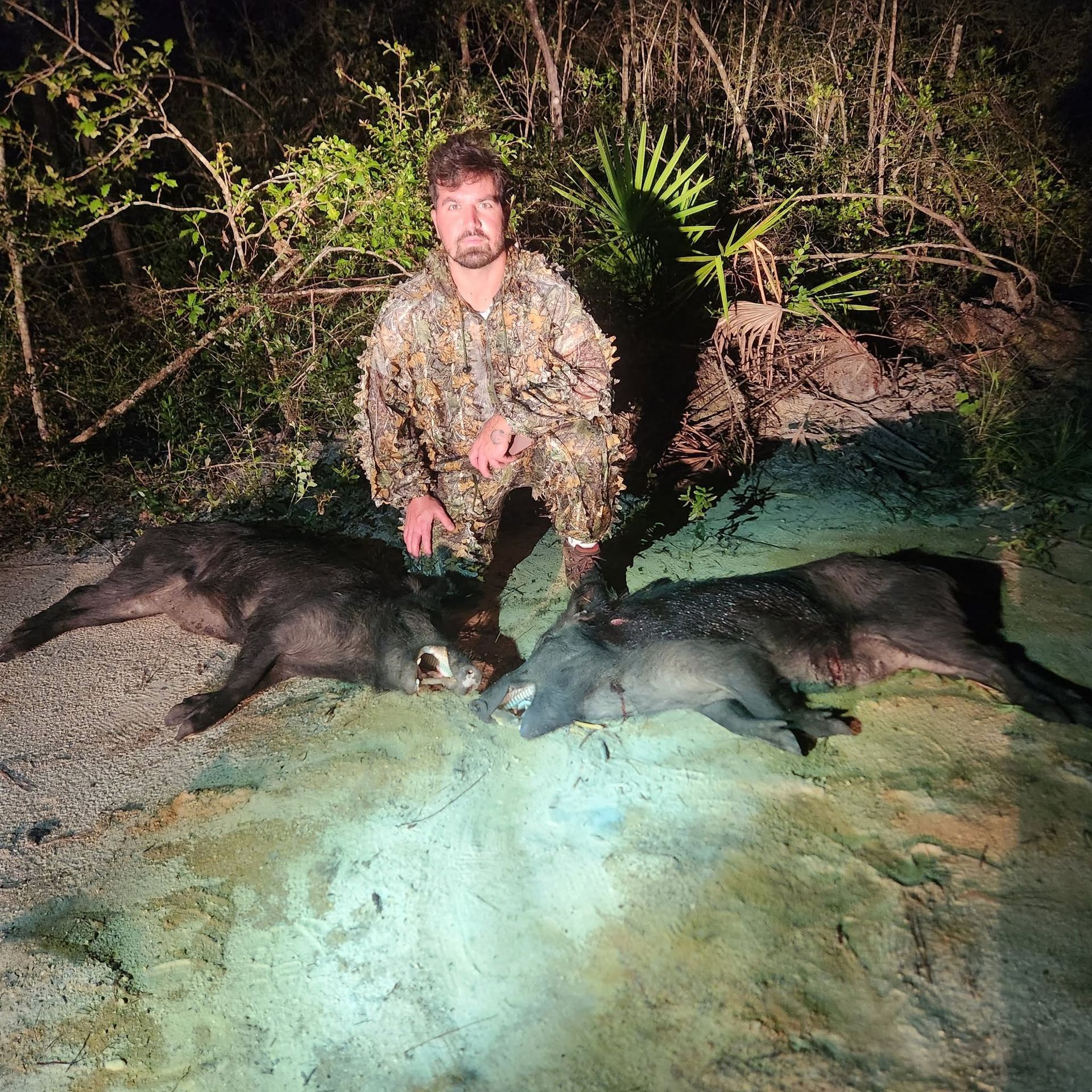 Man crouching beside two dark animals at night, lit by a bright flashlight near water and brush