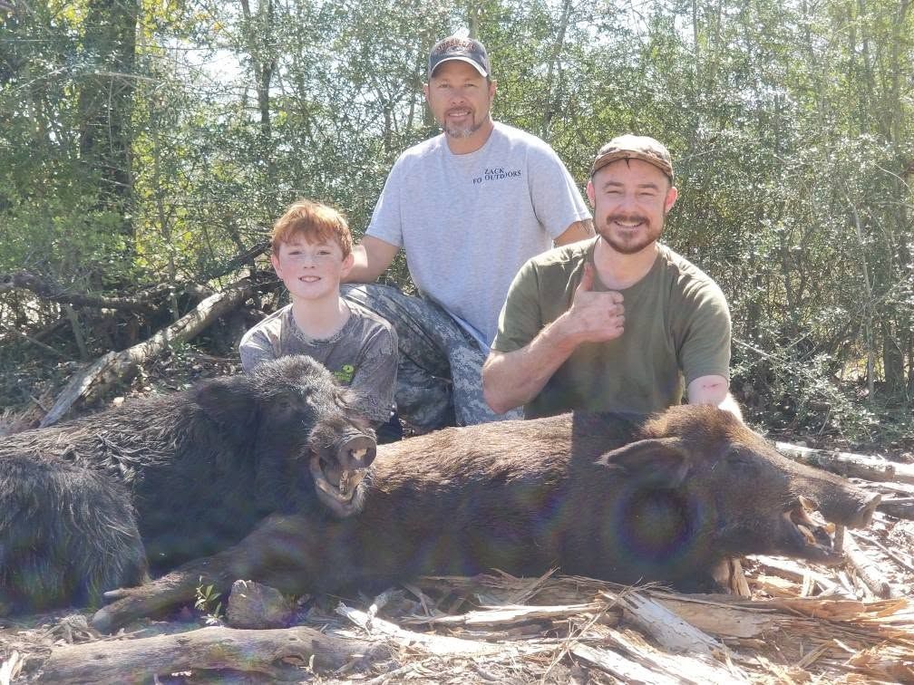 Three hunters posing outdoors with two dead wild boars in front of them.