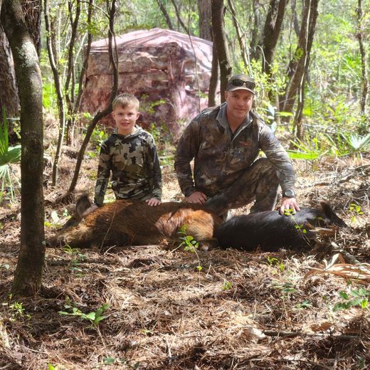 Two people in camouflage posing outdoors beside a large downed log in a wooded area
