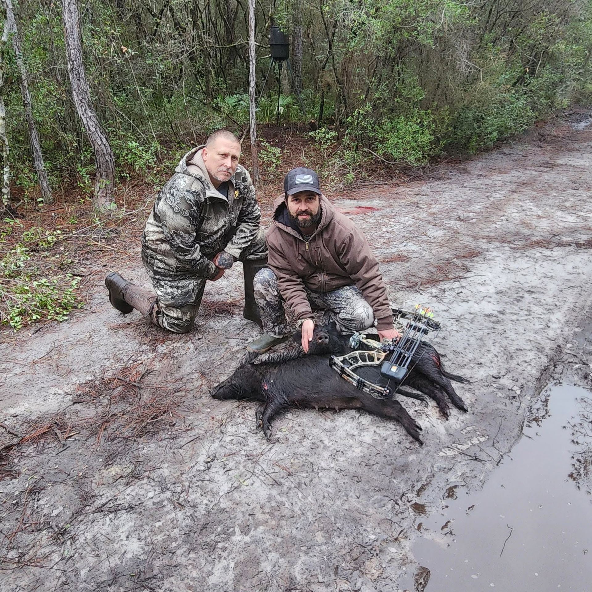 Two hunters kneel beside a wild boar on a muddy forest trail, one holding a device.
