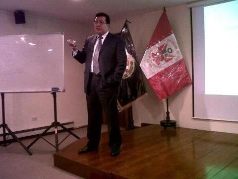 Un hombre habla en un pequeño escenario junto a un rotafolio y banderas peruanas, en una sala de reuniones.