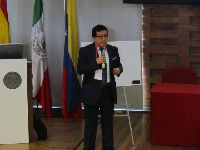 Un hombre con traje negro habla por un micrófono en un podio junto a banderas en una sala de conferencias.