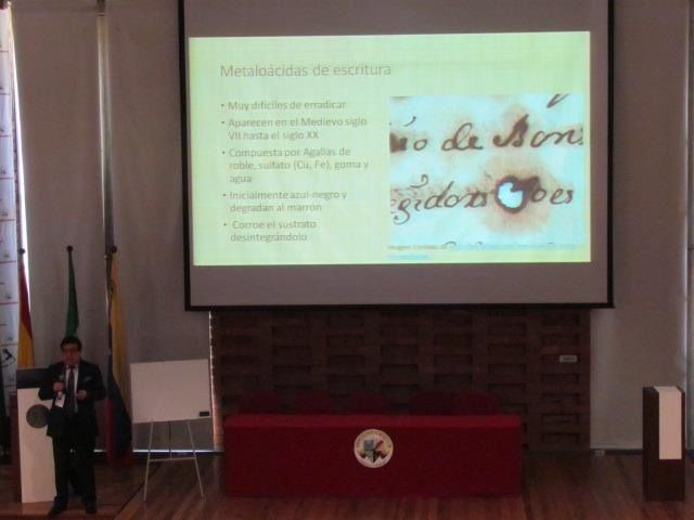 Persona realizando una presentación en un aula junto a una diapositiva proyectada en una pantalla.
