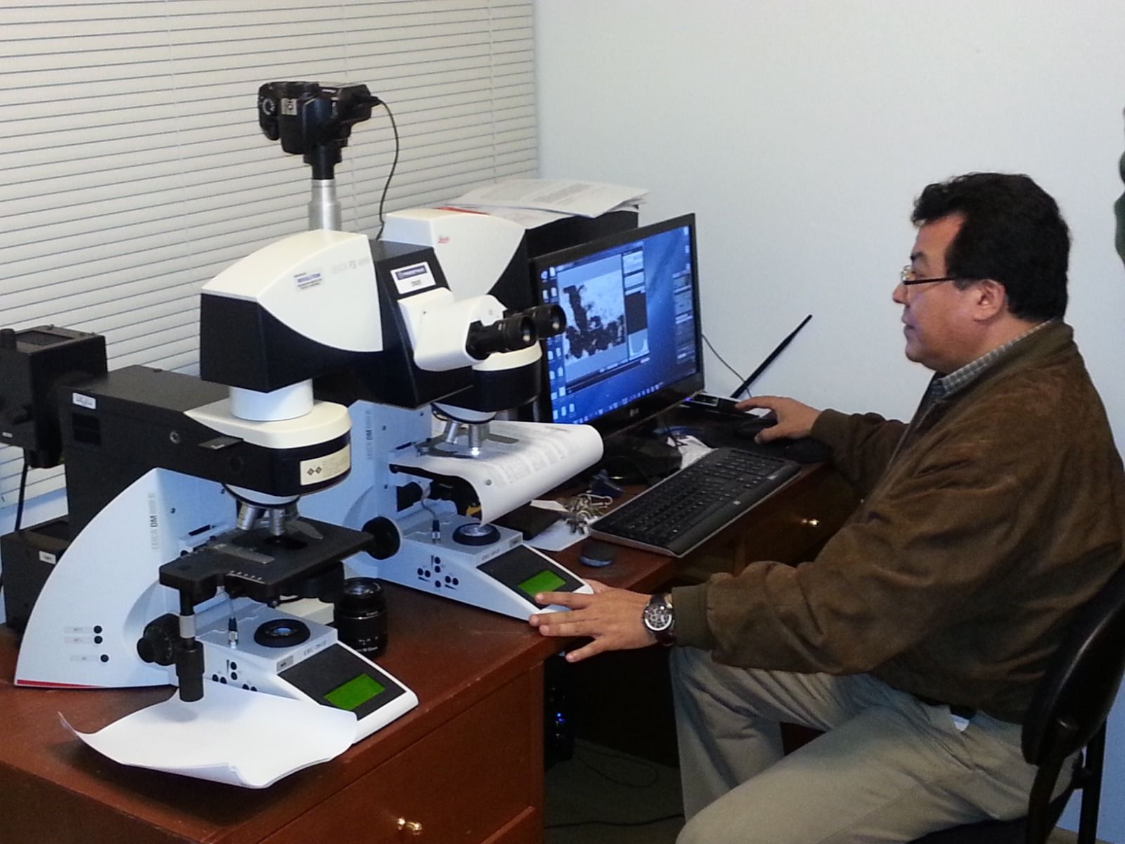 Investigador sentado en una estación de trabajo con microscopio, observando la pantalla de una computadora en un laboratorio.