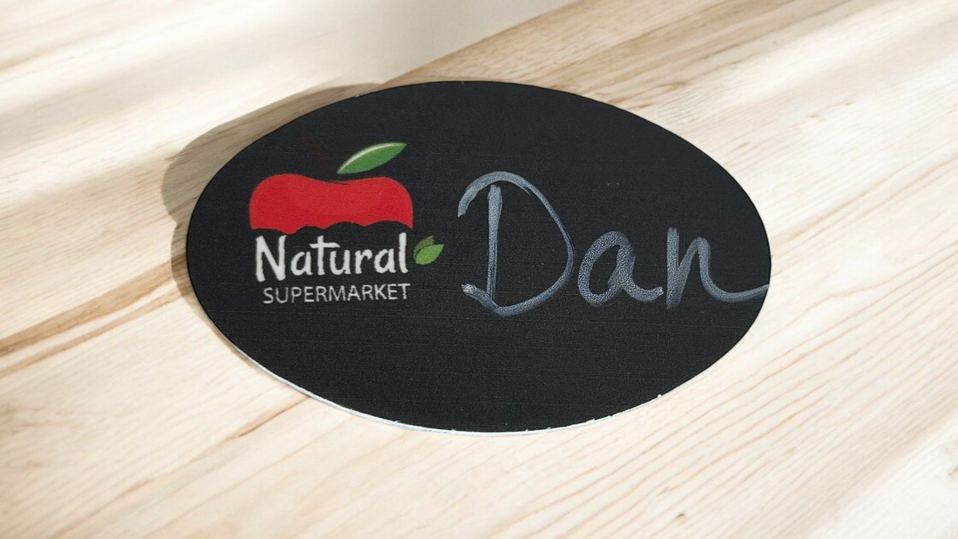 Letrero ovalado negro de “Supermercado Natural” con el logotipo de una manzana roja y el nombre “Dan” escrito con tiza.