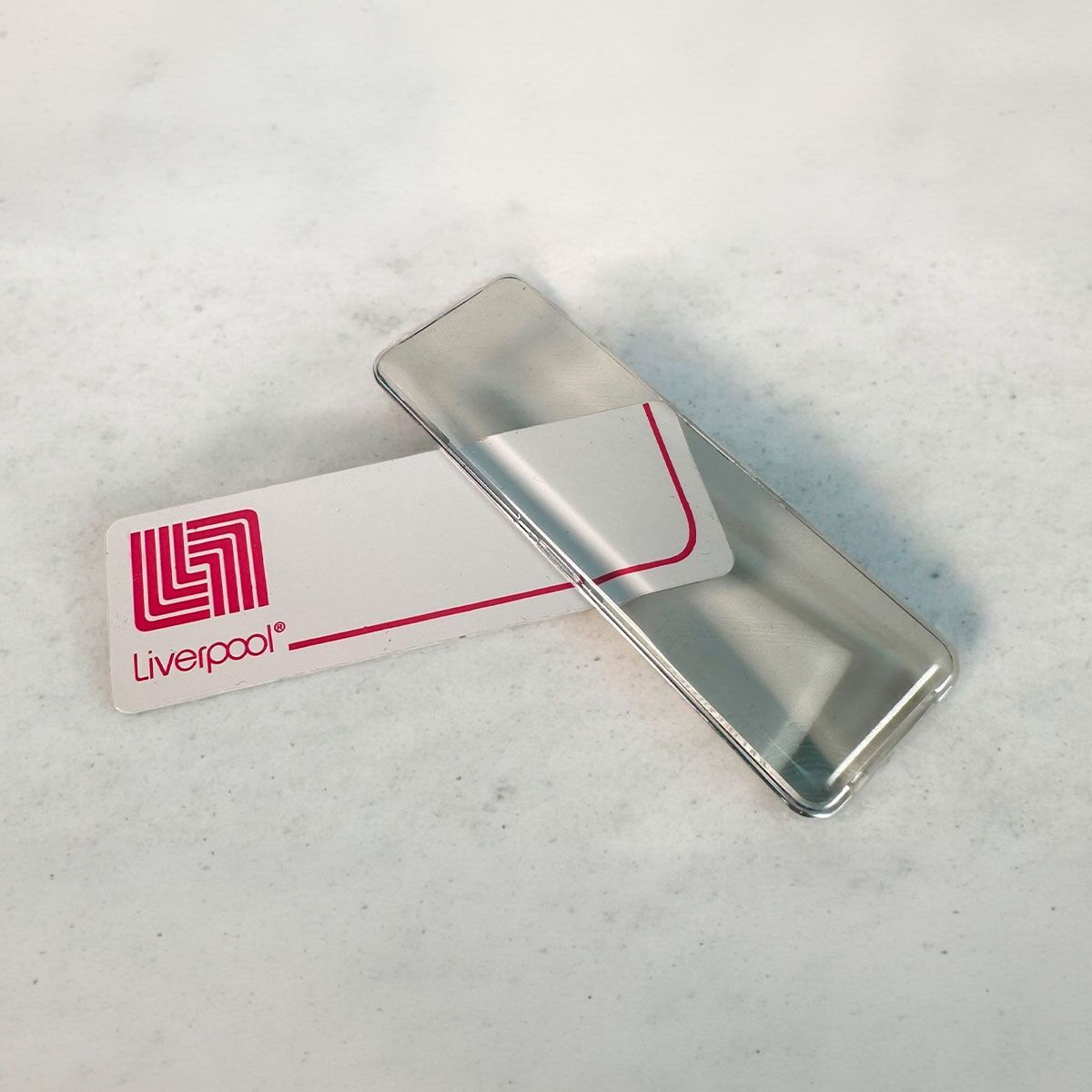 Encendedor de metal plateado junto a una tarjeta con la marca Liverpool sobre una superficie blanca.