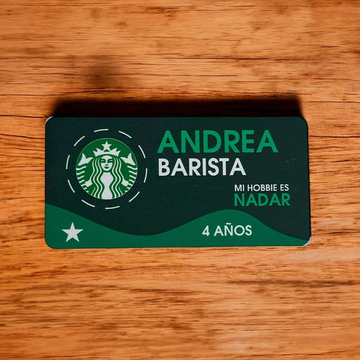 Tarjeta verde estilo Starbucks con “ANDREA BARISTA”, “Mi hobby es nadar” y “4 años” sobre una superficie de madera.