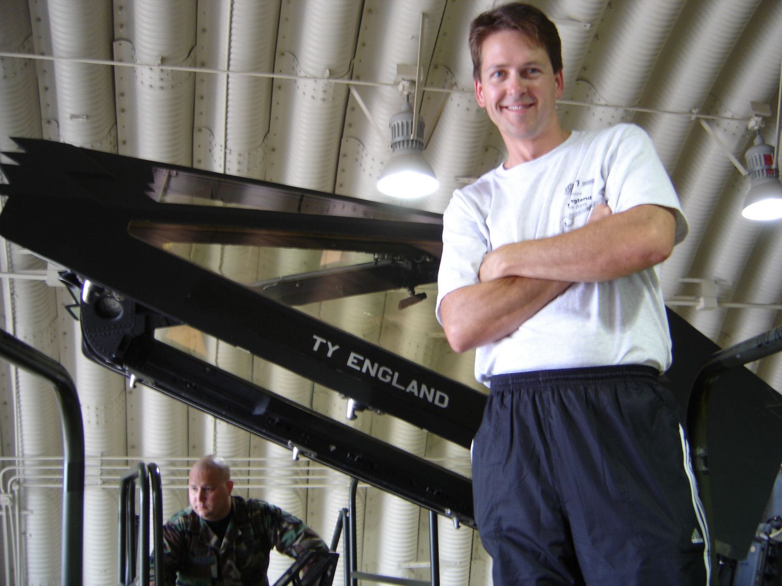 A smiling Ty England stands with arms crossed in front of a military vehicle in a hangar, with a worker nearby.