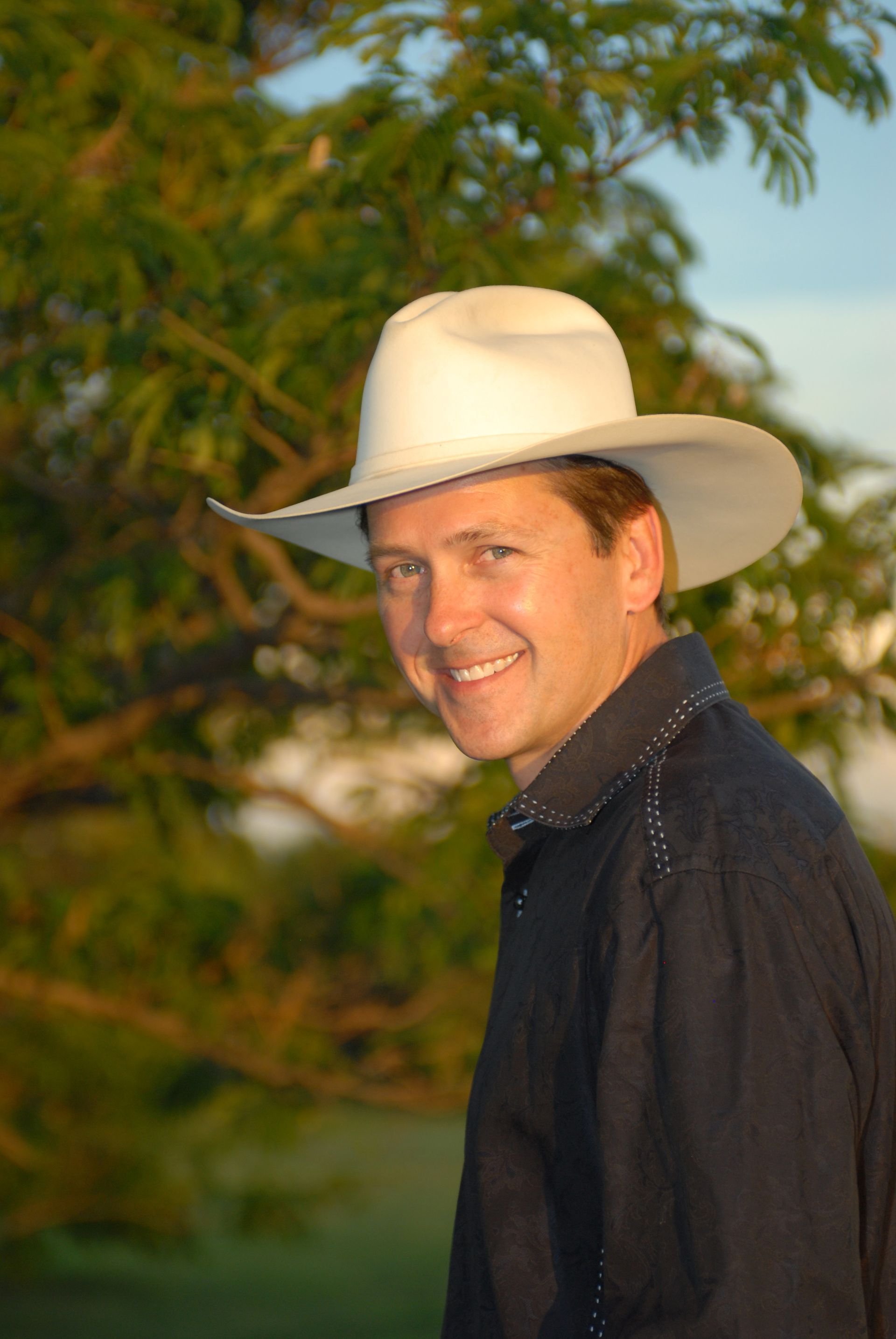 Ty England wearing a white cowboy hat and a dark button-down shirt smiles while outdoors with a blurred tree background.