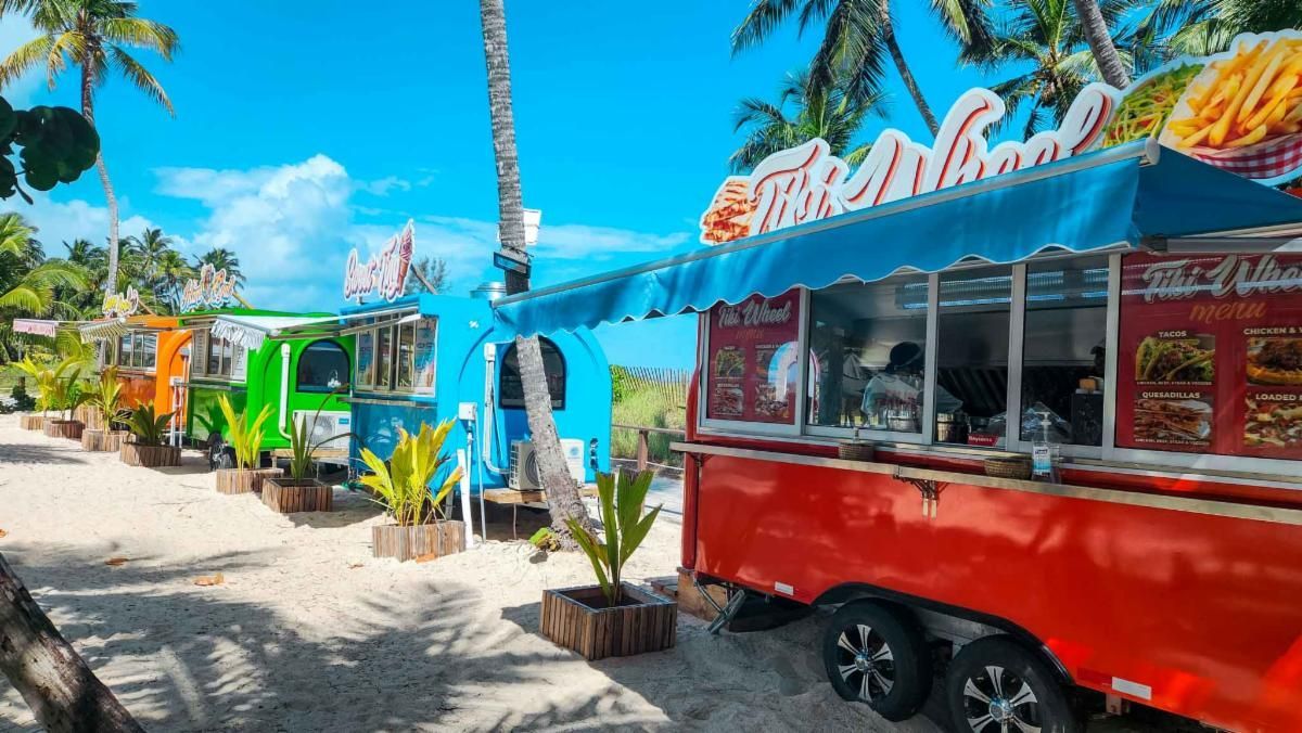 Colorful food trucks lined up on a sandy beach under a bright blue sky. Palm trees are visible in the background.