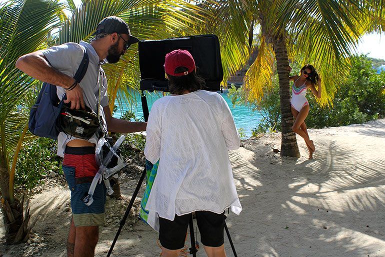 Two people set up equipment for a photo shoot on a tropical beach, with a model posing in a white swimsuit near a palm tree.