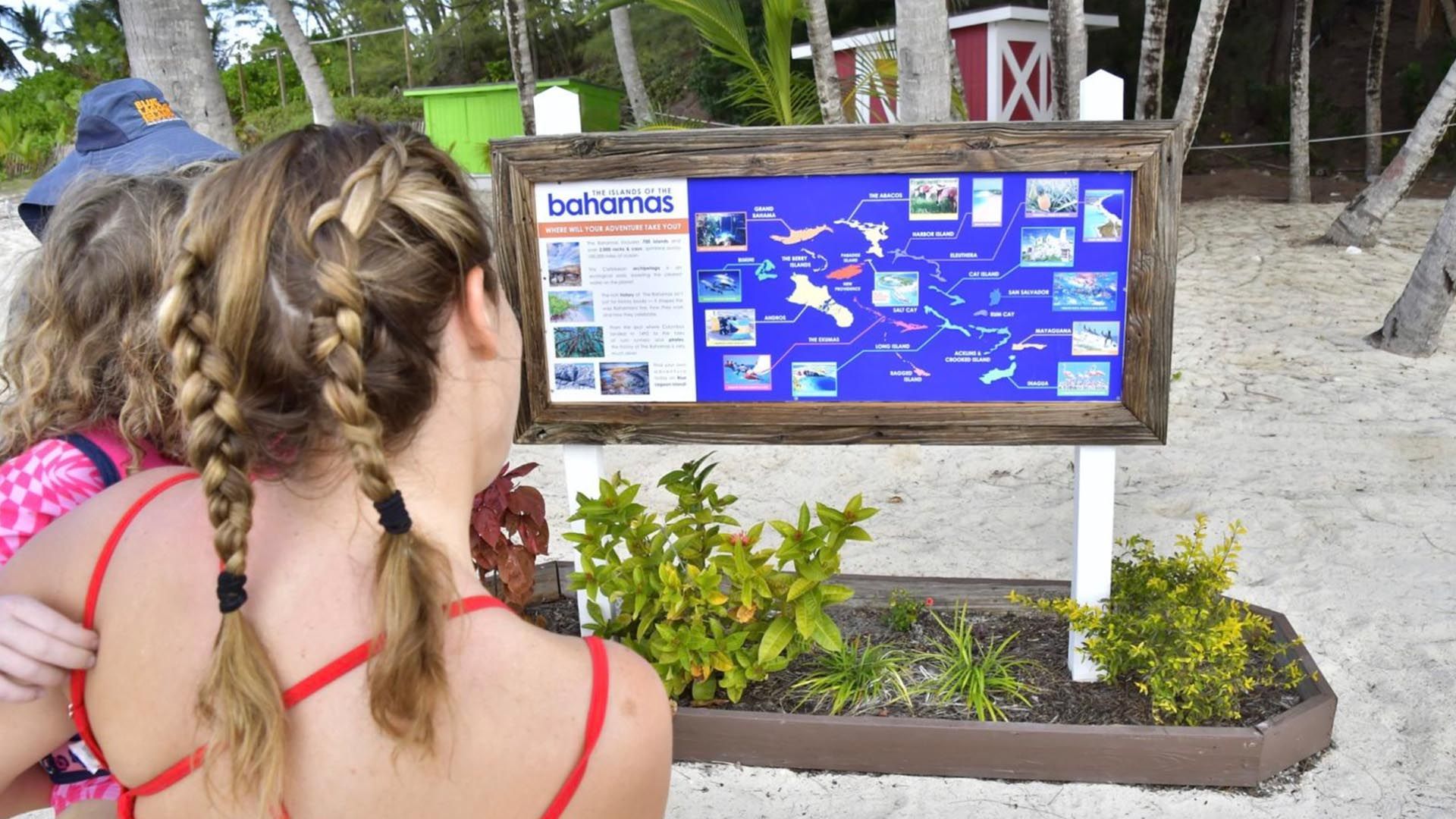 Two women look at a map of The Bahamas on a wooden sign on a beach. One has braids.