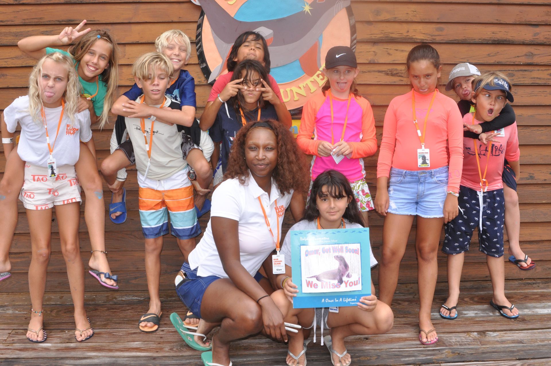 A group of children and two adults smiling at the camera outdoors, some making silly faces, in front of a wooden wall.