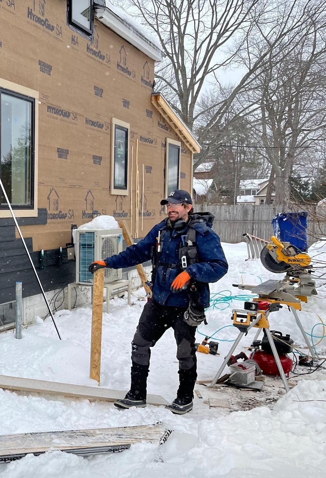 Owner Clint Horsely, stands by a house in snowy yard with tools and equipment nearby.