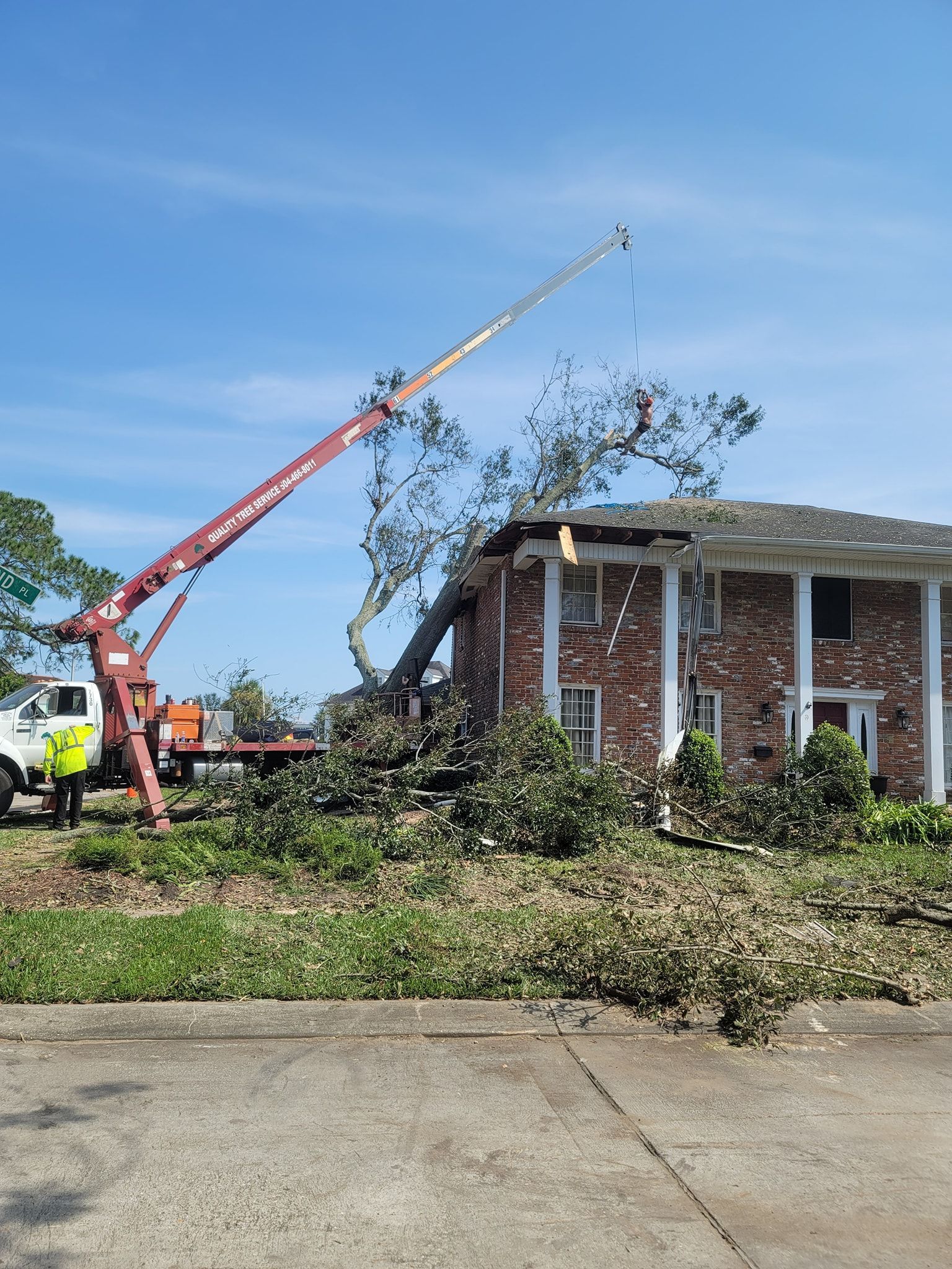 A red crane removes a fallen tree from the roof of a two-story brick house on a sunny day.