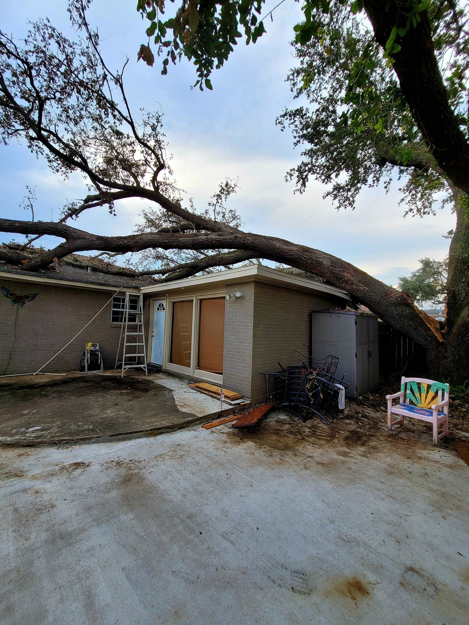 A large tree limb has fallen across the roof of a brick house, partially covering the patio and exterior wall.