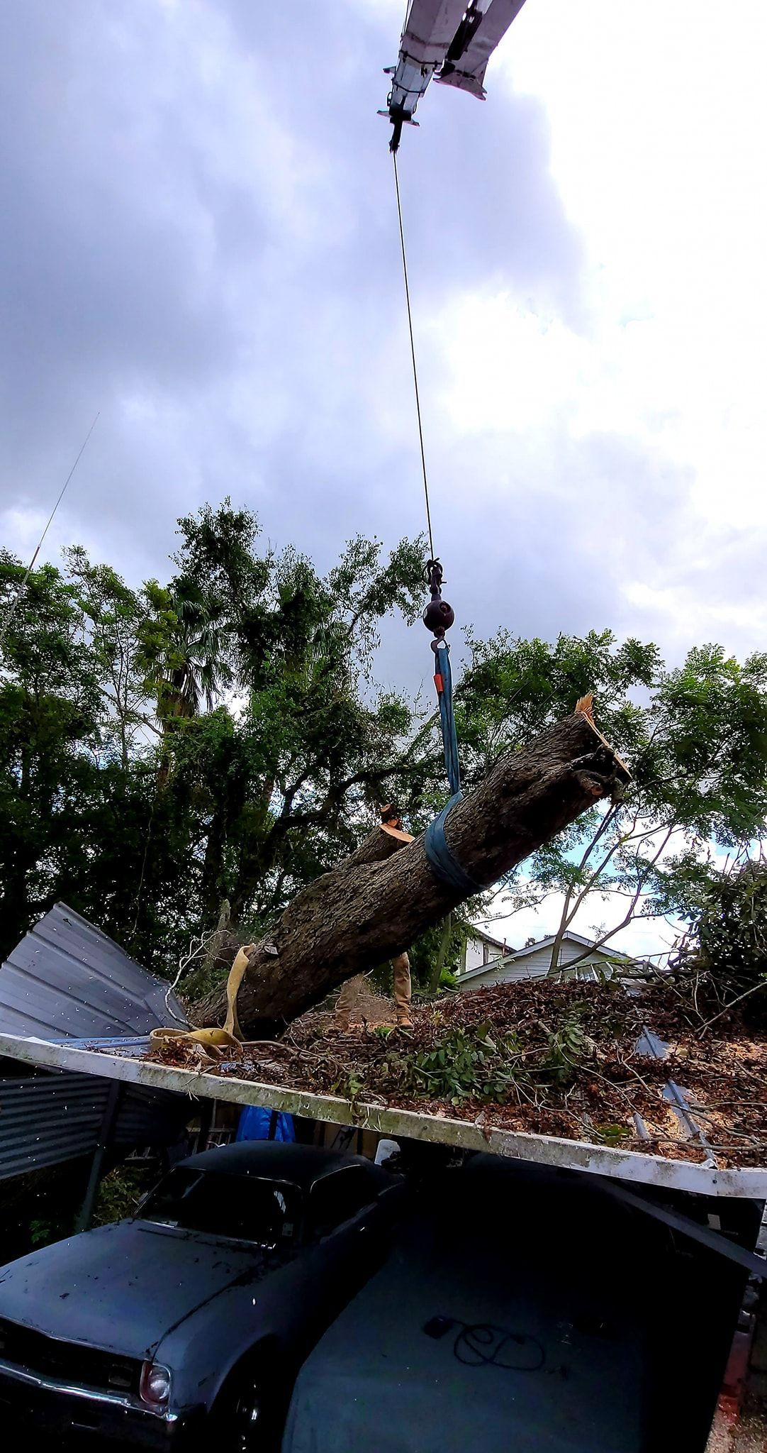 A crane lifts a large tree trunk over a house, with a car parked in the driveway below under a partially damaged roof.