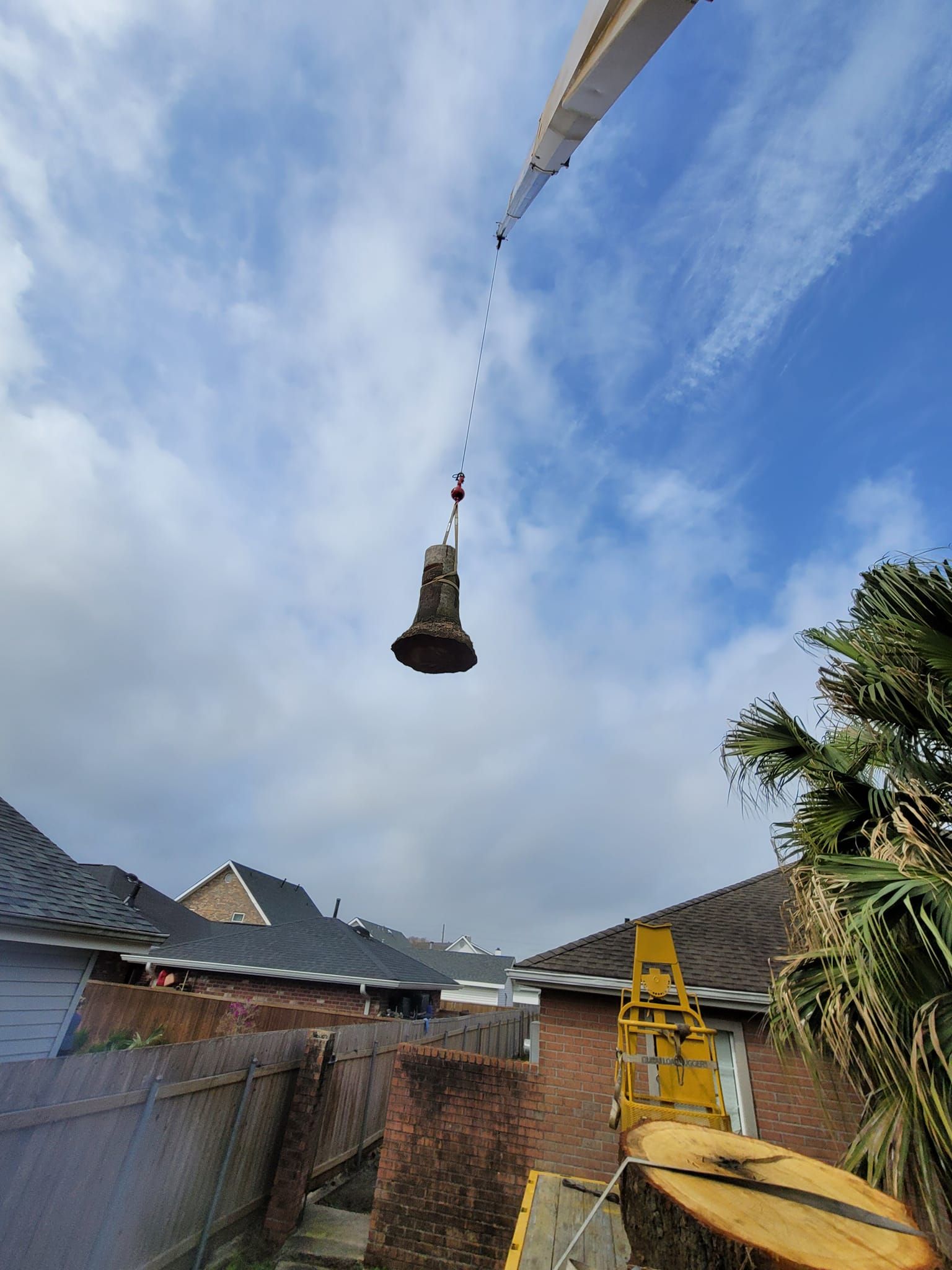 A crane lifts a large tree trunk section over a suburban house and yard under a blue sky.