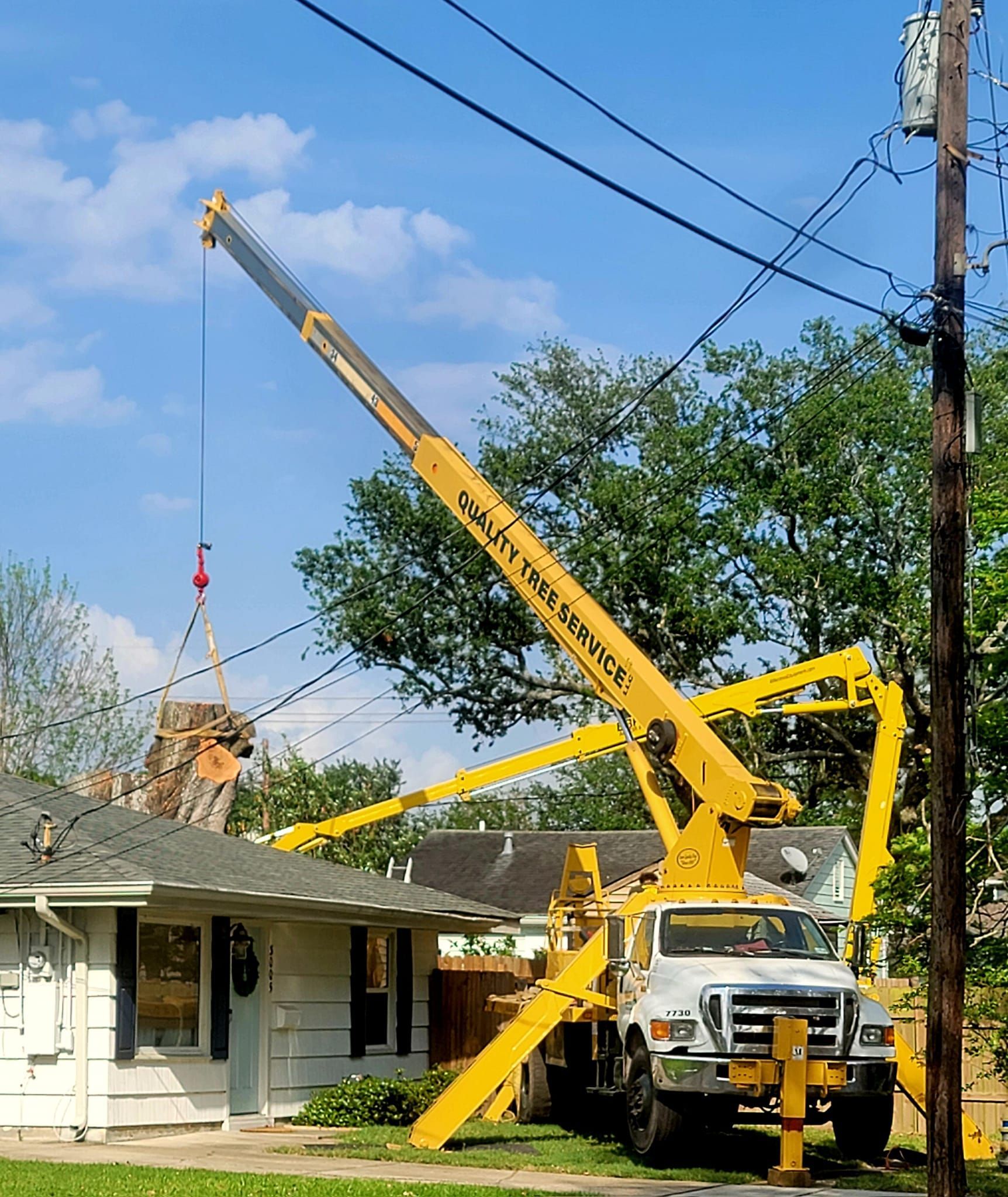 A yellow crane lifts a large tree trunk section over a house roof in a residential area.
