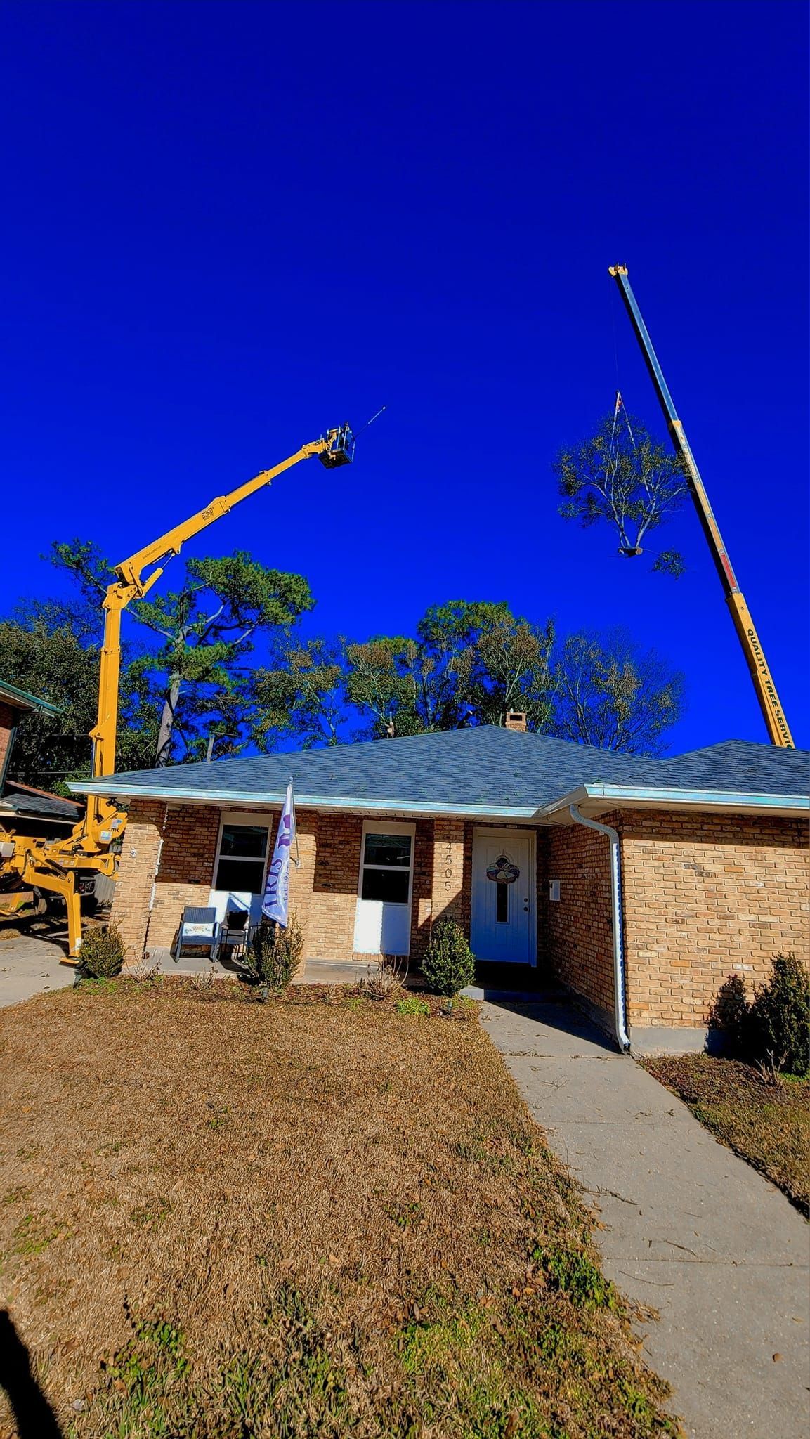 A yellow boom lift reaches toward tree branches above a single-story brick house with a blue roof under a clear blue sky.