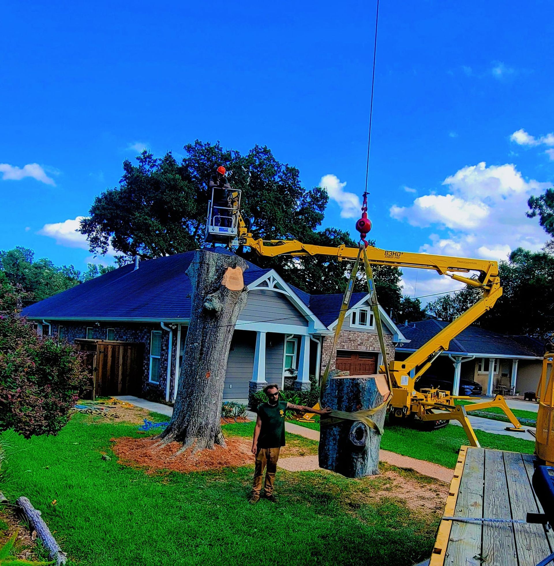 A yellow crane lifts a cut tree trunk section near a house while a worker stands on the lawn nearby.