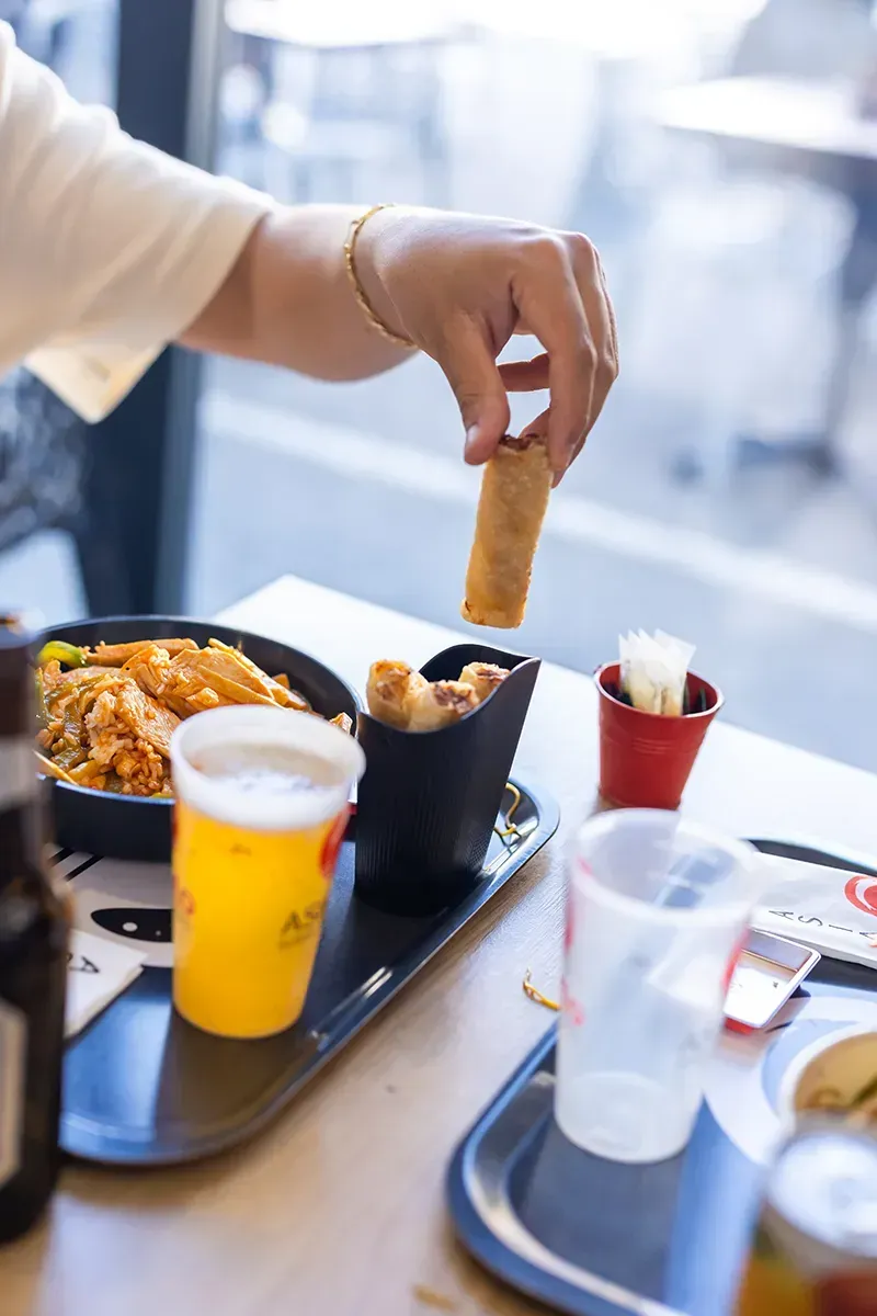 Une personne tenant un rouleau de printemps et le trempant dans de la sauce, à une table de restaurant où se trouvent également de la nourriture, des boissons et des couverts.