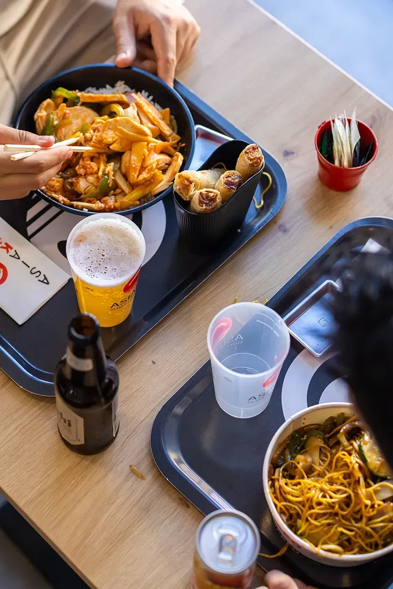 Vue aérienne de deux personnes mangeant des plats asiatiques servis sur des plateaux : nouilles, bière et rouleaux de printemps sur une table en bois.