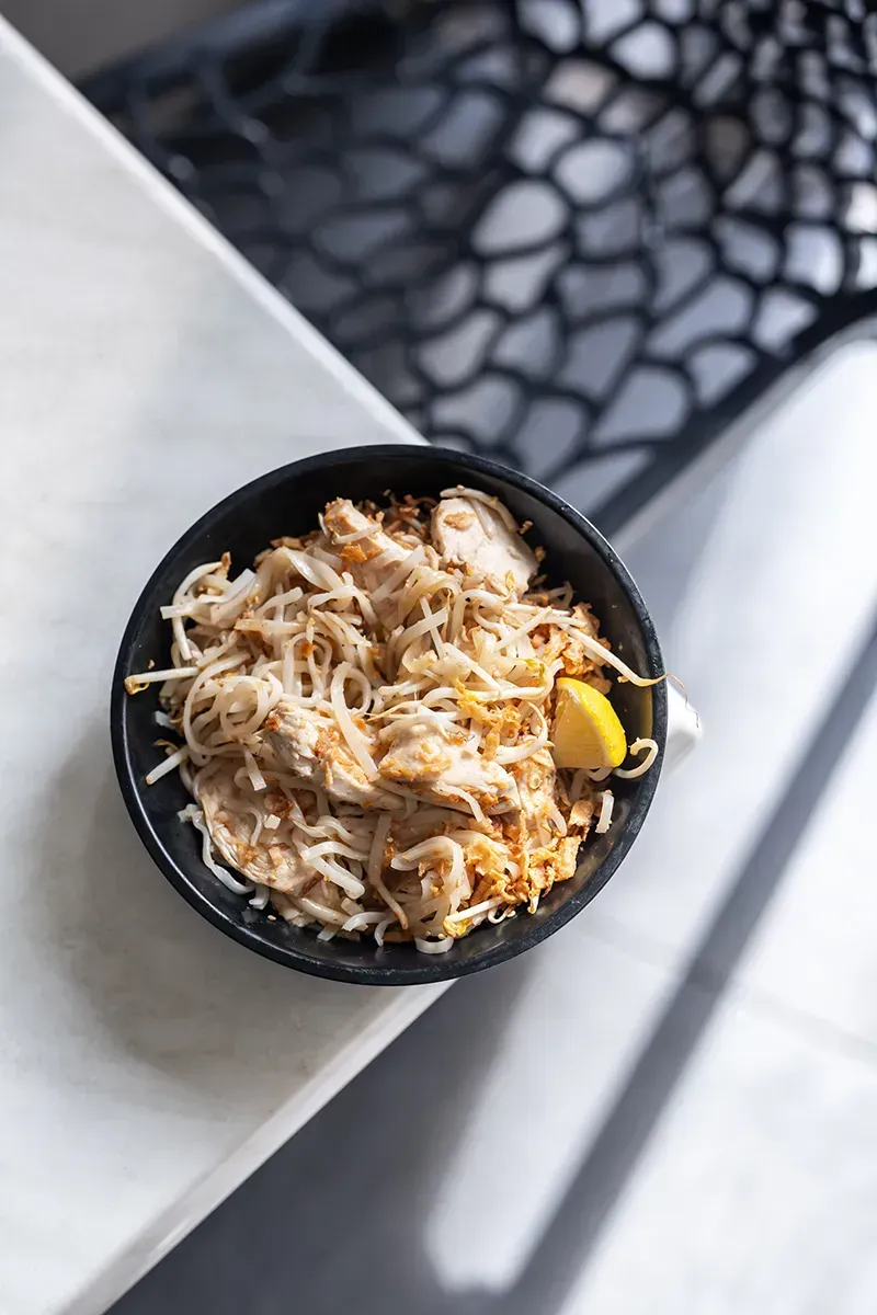 Un bol de nouilles au poulet et un quartier de citron sur une table blanche.
